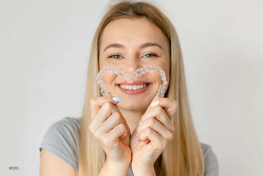 A blonde woman displays her Invisalign® aligners in the shape of a heart.